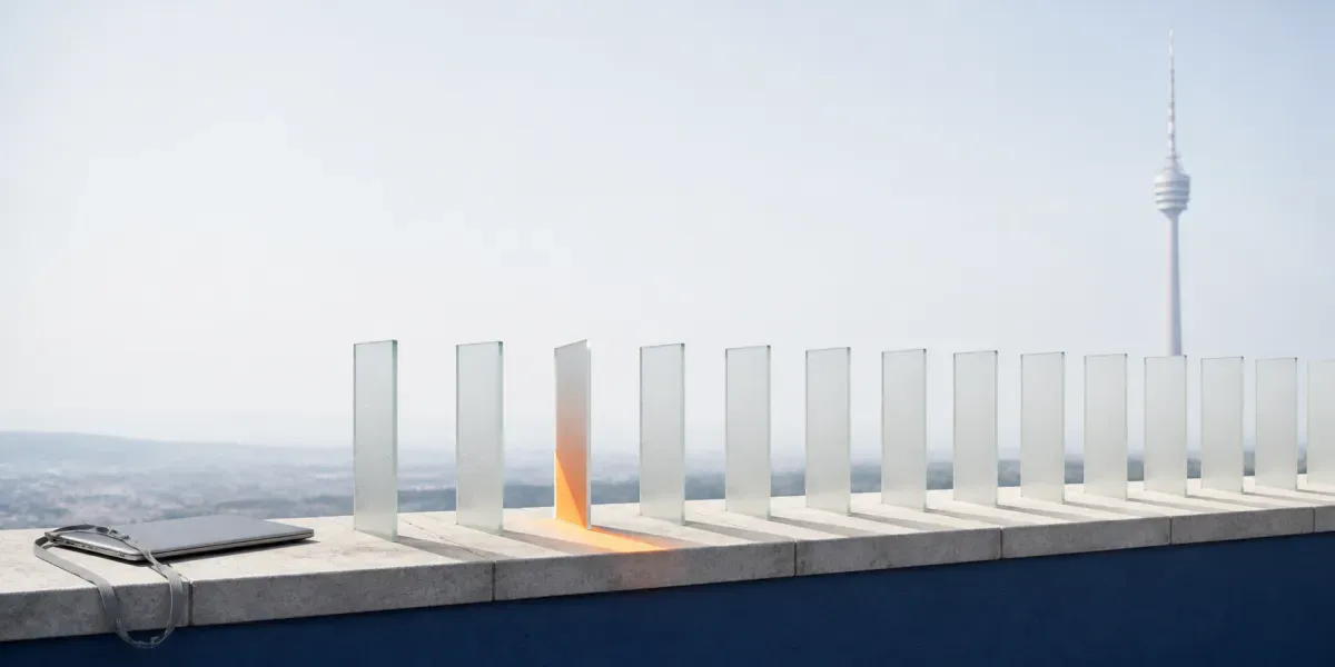 Auf einer Brüstung über Stuttgart steht eine Reihe gleicher Glaslamellen; eine einzige ist anders gedreht und fängt als einzige das warme Morgenlicht ein, im Dunst dahinter der Fernsehturm