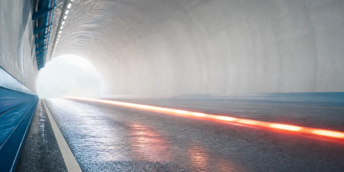 Blick durch einen hellen Straßentunnel auf die taghelle Ausfahrt: eine einzelne orange Lichtspur läuft über die nasse Fahrbahn und verdichtet sich zur Öffnung hin von breiten, unterbrochenen Pulsen zu einer straffen, durchgehenden Linie