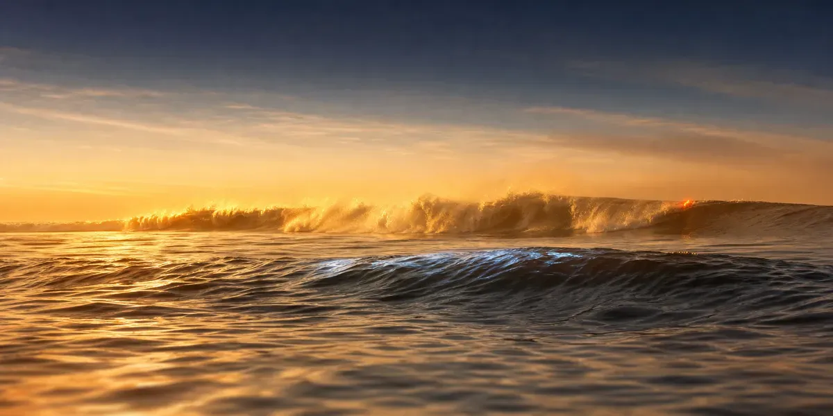 Zwei parallele dunkle Atlantikwellen, die erste mit brechendem Kamm und einem einzelnen orangen Lichtpunkt, dahinter die zweite Welle in tiefem Navy